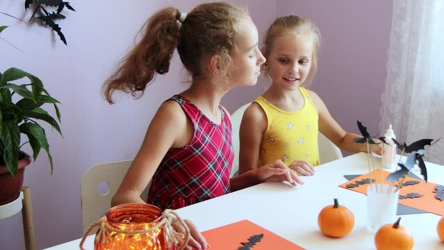 selective focus on two cute blonde schoolgirl sisters, they sit at a table and cut out Halloween symbols from paper, making decorations for the holiday