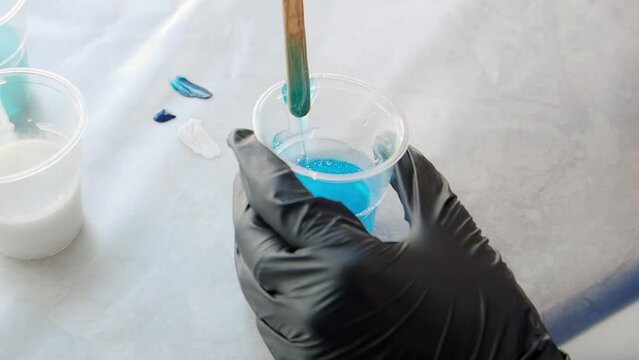Artist mixing white epoxy resin in the plastic cup. Hobby, master class on working with epoxy resin. Interesting pastime. The master stirs the blue paint with a wooden stick.