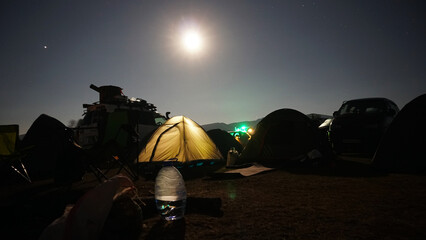 A big bright moon in the starry sky. There is a tent camp. The yellow tent is glowing. Several cars...