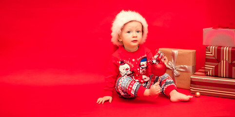 Little Santa with a Christmas tree toy in his hands sits next to Christmas presents on a red background