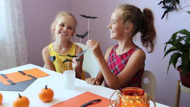 a selective focus of two funny and cute schoolgirls, they are sitting at a table, watching each other and playing with paper bats that were made for Halloween