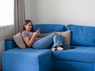 Obraz premium Woman with dark hair in jeans and T-shirt lying on a blue sofa in her living room and reading the news on a phone. Female at home using smartphone for checking social networks or watching films online