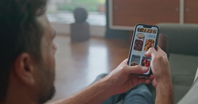 Man orders fast food with phone sitting at living room sofa. Person looking for a good place to eat with smartphone application. Modern technology device. Ordering online food