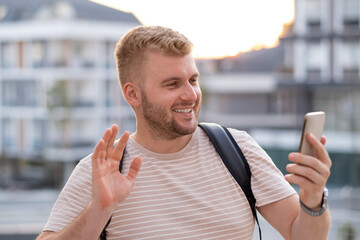 Handsome blond bearded smiling man with casual clothes,having video call using smartphone in city outdoors.Happy caucasian guy waving hand, street photography.Modern technology communication concept.