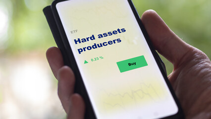 An investor's analyzing the hard assets fund on screen. A phone shows the ETF's prices hard assets producers. 