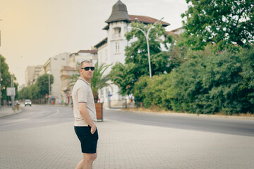 Varna, Bulgaria - 15.08.22 - Young stylish man wearing t-shirt with beard in glasses, standing on the street on city background. Street photo - obrazy, fototapety, plakaty