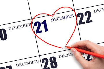A woman's hand drawing a red heart shape on the calendar date of 21 December. Heart as a symbol of love.