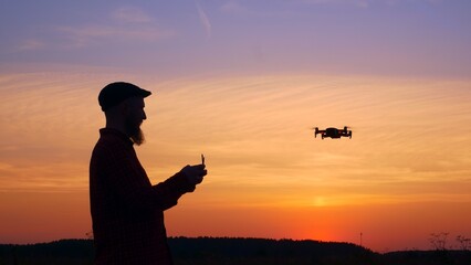 Silhouette of a man with a beard who controls a drone at sunset near the forest. The sky is filled with the beautiful colors of the sunset, which highlights the profile of a man and a drone in the sky