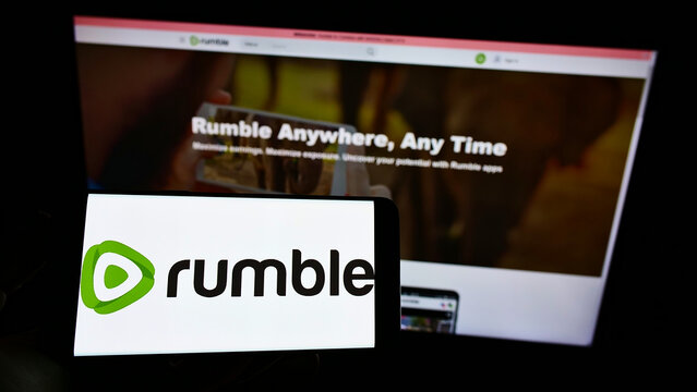 Stuttgart, Germany - 12-19-2021: Person holding mobile phone with logo of Canadian video platform company Rumble Inc. on screen in front of web page. Focus on phone display.