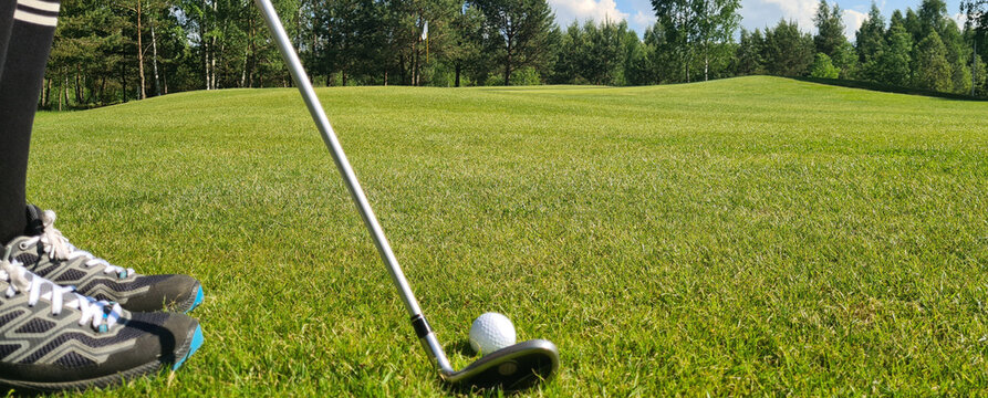 Closeup of white golf ball and club. Human feet on green grass