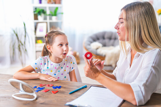 Doctor And Small Patient Train Articulation, Work On Problems And Obstacles Child With Dyslexia. Little Girl Together With Speech Therapist Is Sitting At Desk Indoors, Playing Game, Studying Sounds