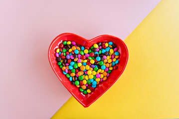 Confetti chocolates on heart-shaped plate on yellow and pink background