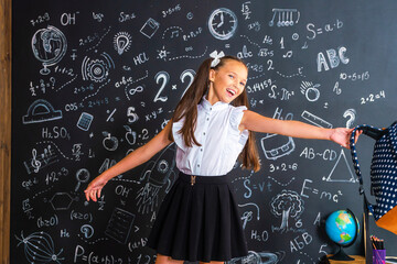 charming mischievous little girl in a school uniform, smiling at the camera and holding a backpack in her hands, has fun during school classes against a black wall. Back to school. Dynamic image