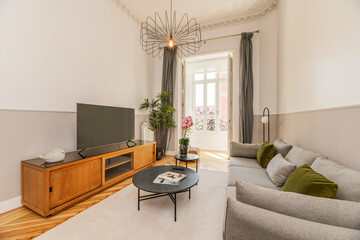 Living room with a three-seater sofa upholstered in gray fabric, a wooden sideboard and a flat-screen TV with a bay window of white windows