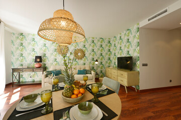 round wooden dining table with designer chairs upholstered in fabric, rattan designer lamp, living room with reddish wood floor and breakfast with fresh fruits and glasses of juice