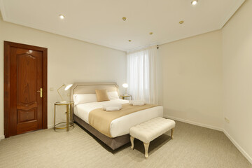 Bedroom with king-size bed with matching cushions and pillows, twin round bedside tables with lamps, upholstered divan at the foot and gray carpet