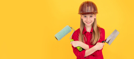 positive teen child in builder hard hat with paint roller and spatula on yellow wall. Child builder in helmet horizontal poster design. Banner header, copy space.