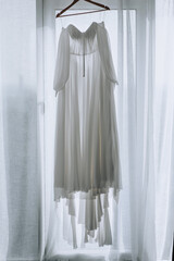 A beautiful long white bridesmaid dress hangs on a hanger in front of the window. wedding photography.