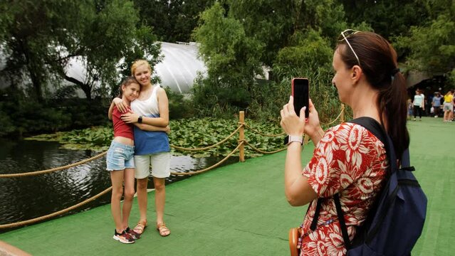 A woman takes pictures of her daughters on her smartphone while walking in the park.