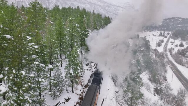 Aerial old steam locomotive, engine rides on railroad releases thick clouds of steam from pipe. Winter snowy nature. Historical American railway in Colorado mountains. Drone flying through smoke