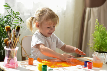 A two-year-old girl sits at a table and gets her hands dirty with paint. Development of fine motor...
