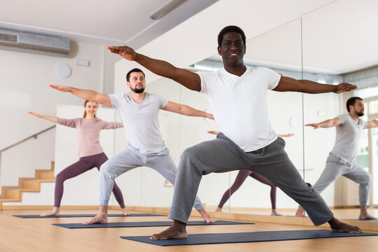 Sporty adult African American doing power yoga with group in fitness studio, standing in lunging asana Virabhadrasana..