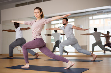 Woman practising warrior II pose with two men during yoga training.