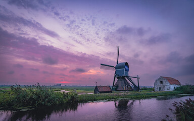 Stary wiatrak i stara zabudowa nad kanałem. Piękne niebo zabarwione przez zachodzące słońce. Wiejski, idylliczny holenderski krajobraz., wiatrak