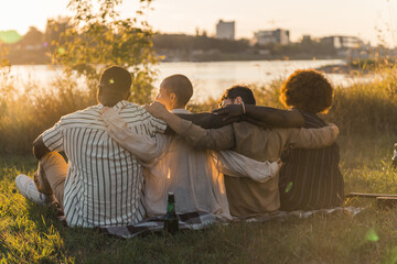 Rear view of multicultural good old friends hugging while sitting on a blanket on the grass, having...