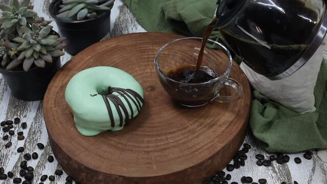 Pouring coffee into transparent cup with donut on the side. Wood, fabric and plants background in green tones