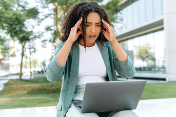 Disappointment puzzled brazilian or hispanic young business woman, elegantly dressed, sit outdoors with laptop, looking at screen in confusion, received unexpected news, holds her hands on her head