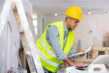 Man engineer checking documents and using laptop computer during repair works indoors.