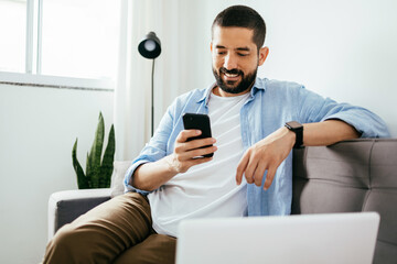 Smiling man sitting on couch at home using laptop and cell phone - Powered by Adobe