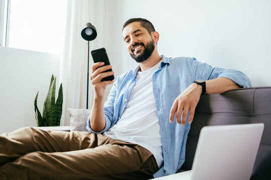 Smiling man sitting on couch at home using laptop and cell phone