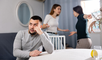 Portrait of sad young adult man sitting at table ignoring conflict of two women at home