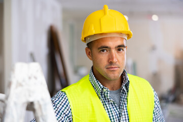 Portrait of man builder in yellow vest and helmet standing in apartment during repair works.