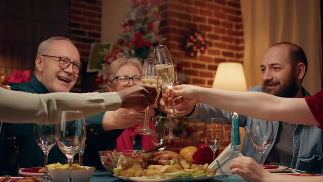 Happy family toasting sparkling wine at Christmas dinner table in living room. Multiethnic positive people gathered inside Xmas decorated apartment with cozy setting to celebrate winter holiday.