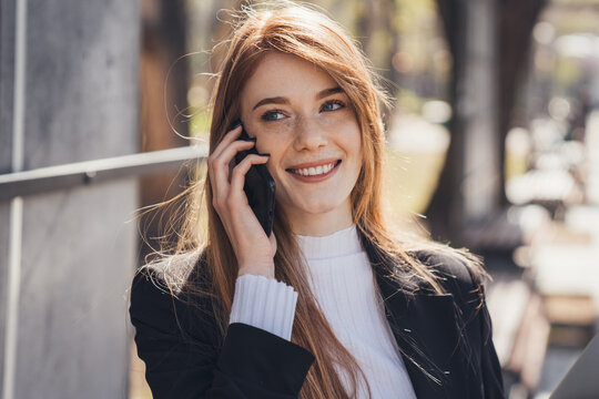 Portrait of a red-haired business woman smiling talking on phone while standing outdoors in the city park. Positive person. Business people communication. Smart