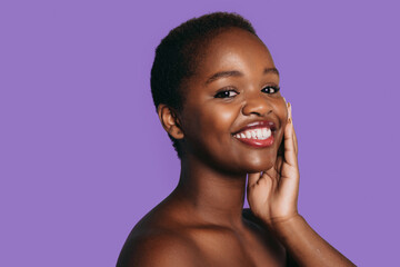 Portrait of attractive African girl touching clean cheeks with perfect glow, isolated on purple background with free space for text. Smiling and looking at