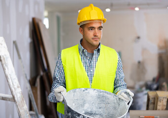 Man builder working in construction site, carrying bucket. Male repairer in yellow vest and helmet lifting bucket.