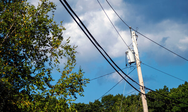 Utility pole with power lines and transformer set against blue sky with dramatic clouds and late summer trees. 