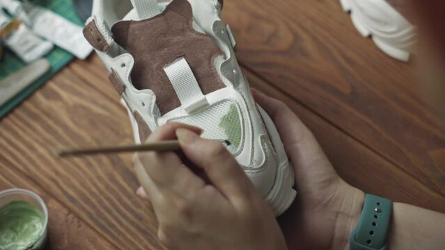 Artist applies green acrylic paint on white sneaker with brush. Tubes of paint and wooden table in background. Hobbies, handmade, crafts and leisure at quarantine.