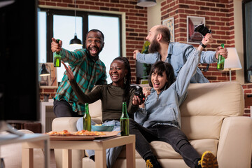 Happy friends playing video games and celebrating victory with beer bottles and pizza at fun gathering. Enjoying hangout at house party with group of people, leisure activity and win.