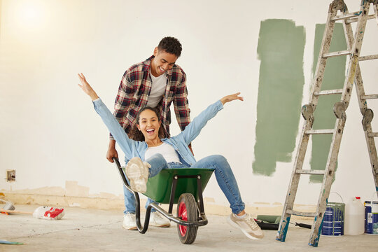DIY, painting and happy couple smile while moving into new home, real estate property house or building. Love, creative man and woman on wheelbarrow interior design and renovation work on room wall