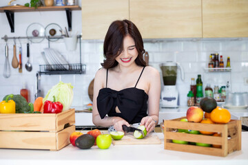 beautiful asian woman smiling preparing, cut green apple in white kitchen with plenty of fresh vegetables and fruits on the table.