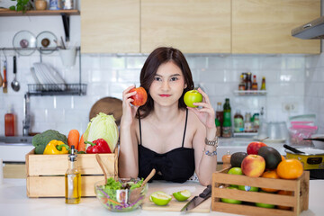 beautiful asian woman smiling preparing, holding a red apple and cut green apple in white kitchen with plenty of fresh vegetables and fruits on the table. At Home