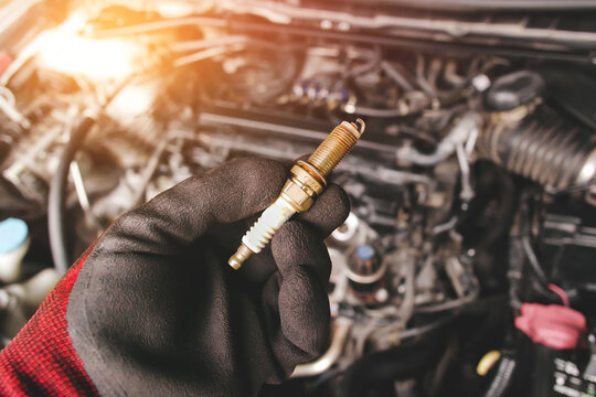 Automobile iridium spark plugs holds by auto mechanic hand with engine compartment blurred background and sunlight.