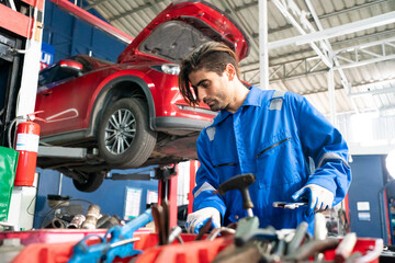 Male car operator wearing blue overalls, gloves working under the hood of car and checking attentively serviceability of engine at repair garage. Concept of car maintenance