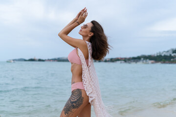 Naklejka premium Stylish young woman with voluminous hair in a bikini and lace pareo posing on a tropical beach, slim, tanned, athletic, sunglasses
