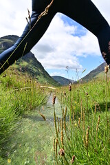 Frau mit langer Jeans macht einen Schritt über einen kleinen Bach, Tal Glen Coe in den Schottischen Highlands, Glencoe, Argyll, Schottland - obrazy, fototapety, plakaty